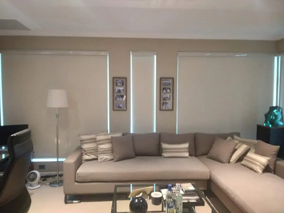 An image of a roller blind made from a white fabric material, covering a window. The roller blind is fully extended and mounted inside the window frame. The bottom of the blind is straight and even, with a thin metal bar that adds weight and stability to the fabric. The fabric is opaque, blocking out most of the natural light and providing privacy for the room. The cord mechanism used to raise and lower the blind is visible on the right-hand side, with the cords hanging down in a neat loop. The roller blind has a sleek and minimalist design that blends in with the window frame and walls, creating a clean and modern look