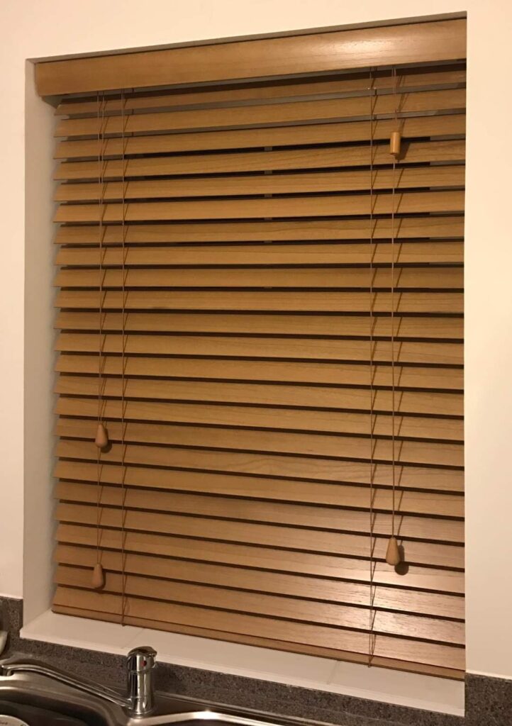 An image of a wooden blind covering a window. The slats of the blind are made from a light-colored wood and are evenly spaced, allowing natural light to filter into the room. The wooden slats are horizontal and can be adjusted to control the amount of light and privacy in the room. The wooden blind is mounted inside the window frame and is shown partially drawn, revealing a view of the outside. The cord mechanism used to raise and lower the blind and adjust the angle of the slats is visible on the right-hand side, with the cords hanging down in a neat loop. The wooden blind adds warmth and texture to the space and complements the other wooden elements in the room, such as furniture or flooring.